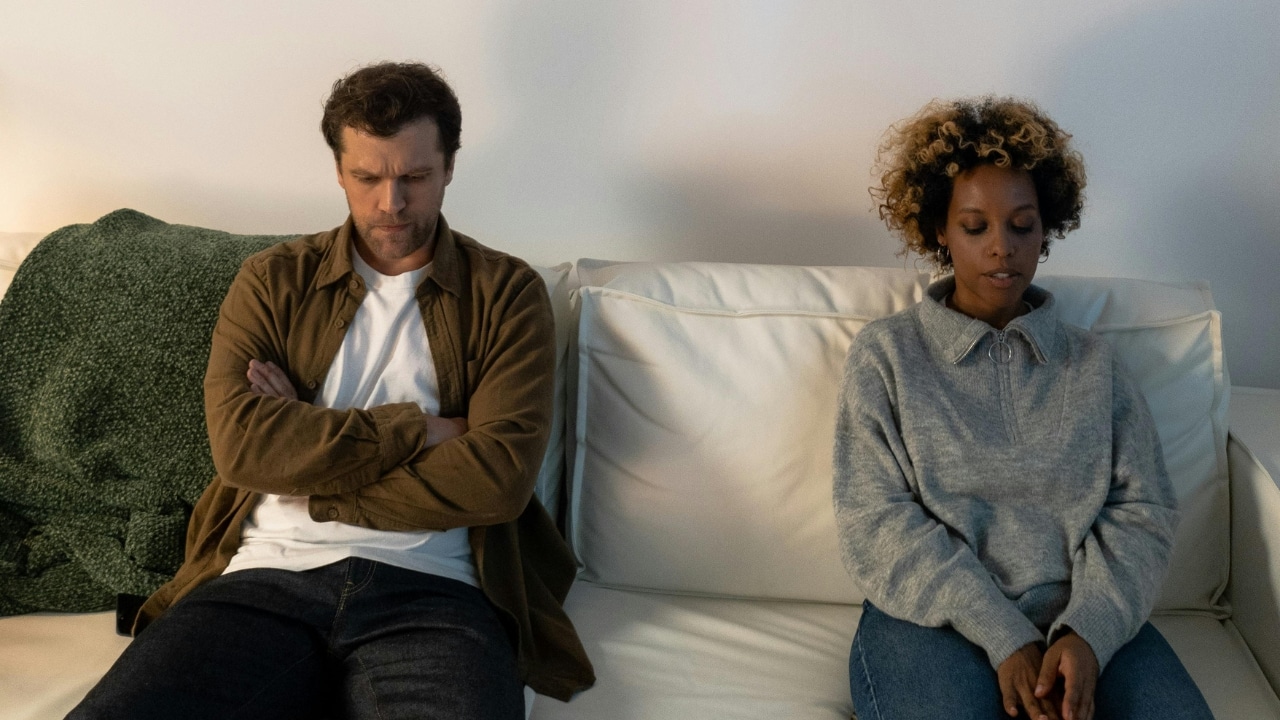 A man with crossed arms and a woman sit apart on a white fabric sofa.