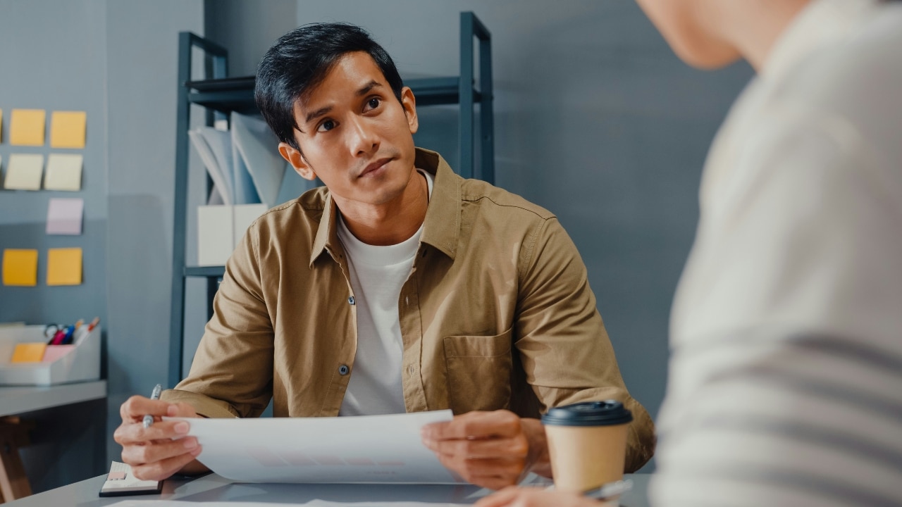 A man in a tan shirt sits at a desk holding papers and looking forward.