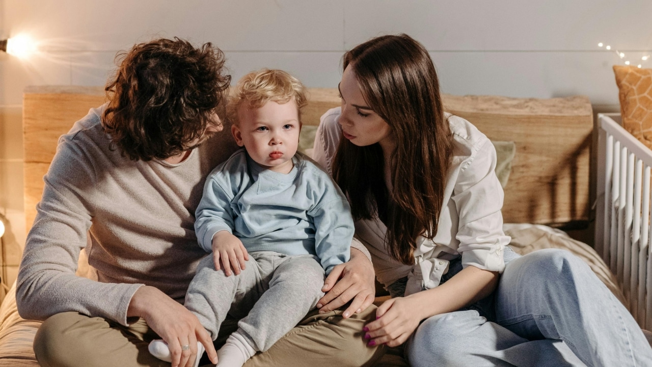 Two adults sit on a bed while looking at a toddler seated between them.