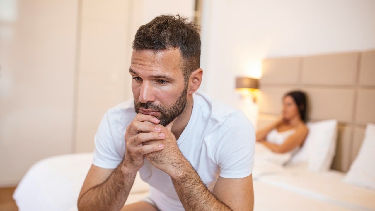 A man sits on a bed with his chin on his hands, looking down thoughtfully.