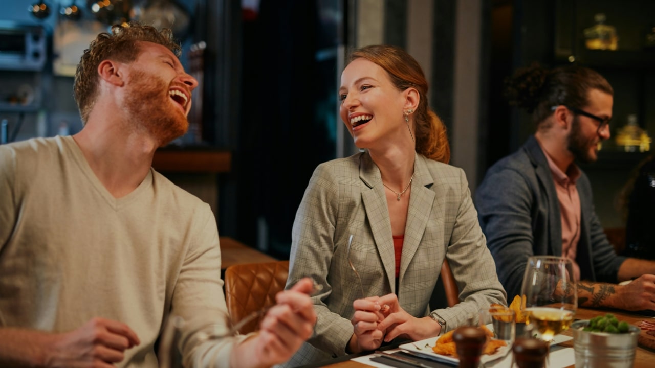 A man and woman laugh while sitting at a dinner table with another man nearby.