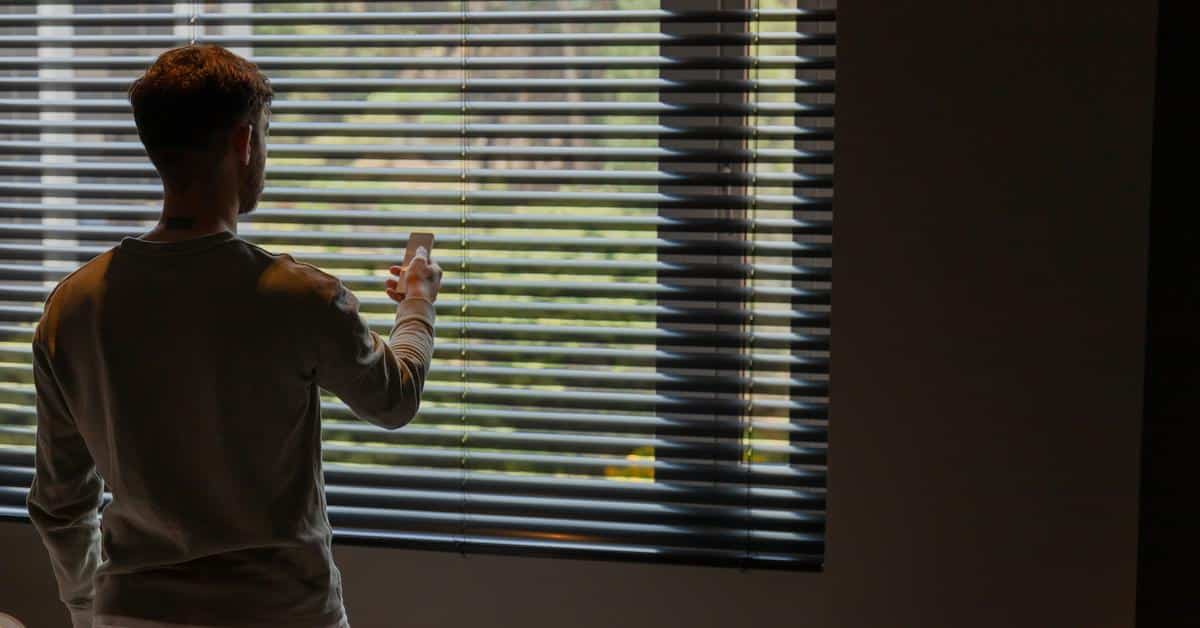 A man standing by window blinds while checking his phone.