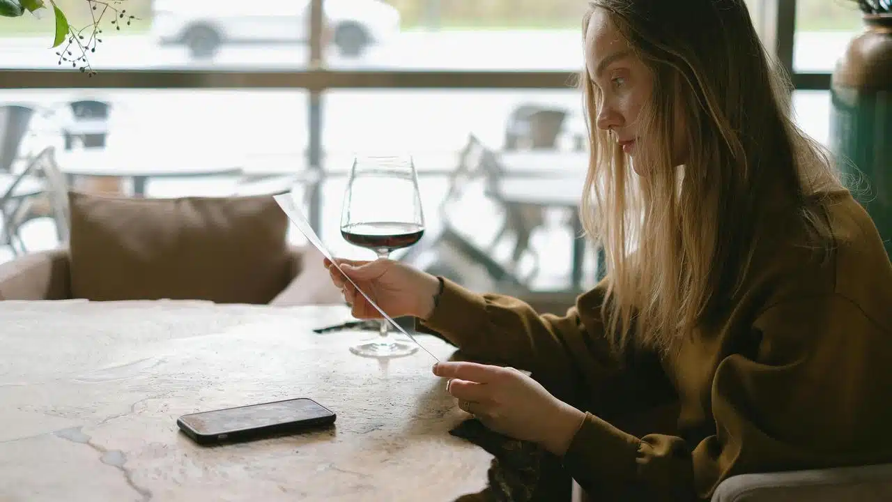 A woman sitting at a table reading a note with a glass of wine nearby.