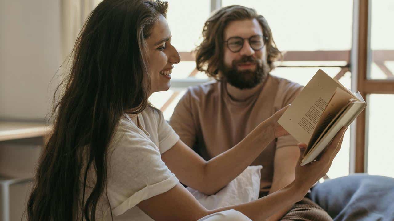 A woman smiling while showing a book to a man sitting beside her.
