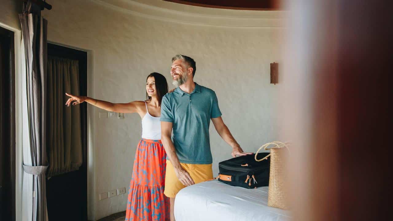 A couple stands in a bright hotel room, smiling and looking out the window.