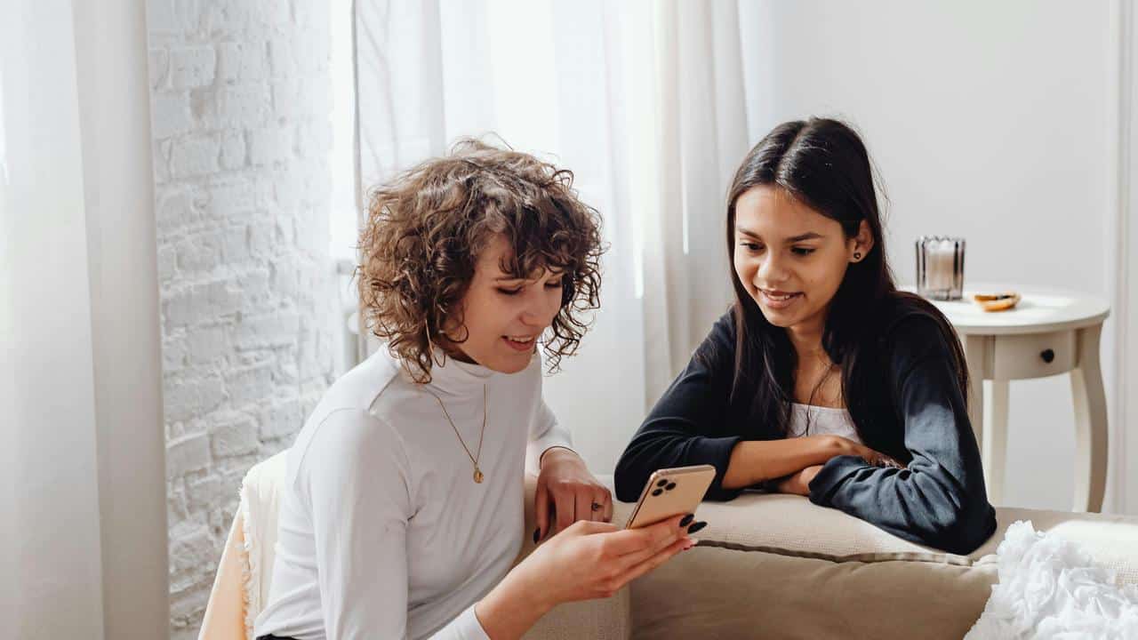 A woman and a girl smiling at a phone together on a couch.
