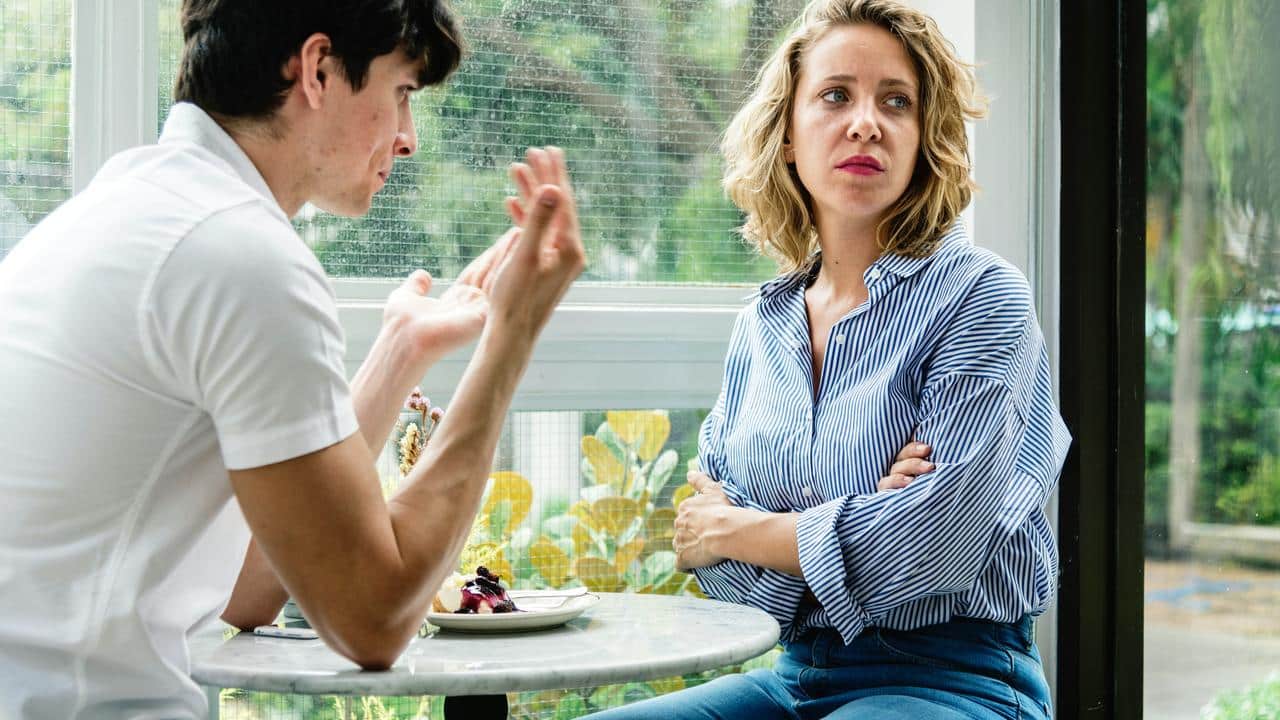 A couple sitting at a table as one gestures while the other looks away with crossed arms.