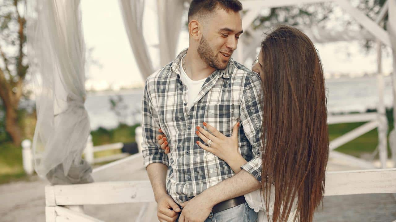 A couple smiling and embracing outdoors near a waterfront.