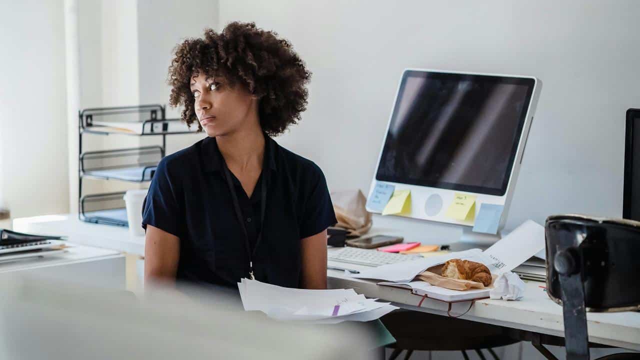 A woman sitting at a cluttered desk in an office, looking away thoughtfully.
