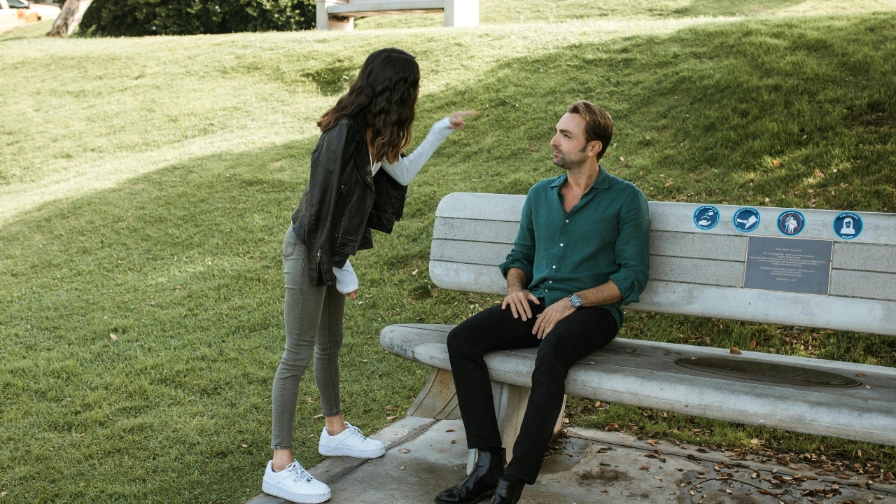 A woman in a leather jacket stands outdoors and points at a man sitting on a bench.