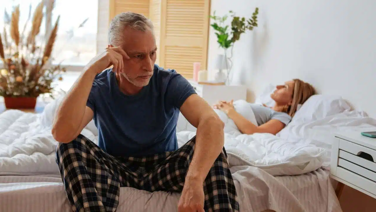 A mature man sits on a bed, looking troubled, with a woman sleeping behind him.