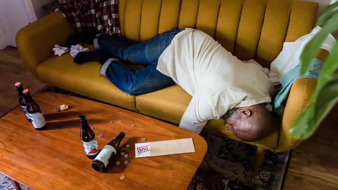 A man lies face down on a yellow sofa next to a table with beer bottles.