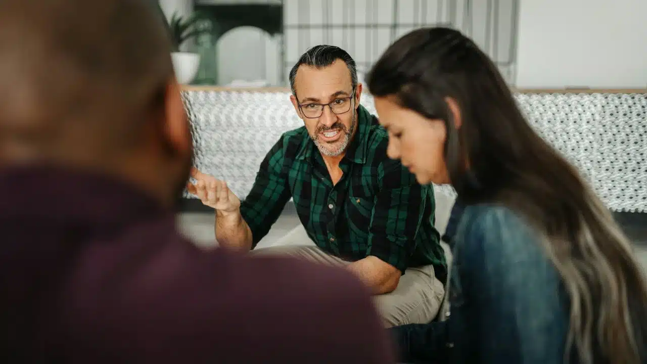 A man in a green plaid shirt speaks animatedly to two people in a room.