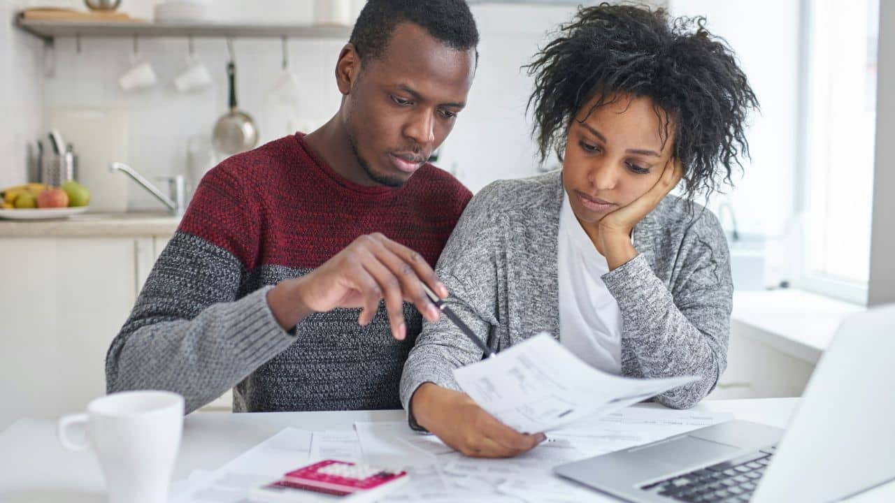 A man and woman sit at a table looking at papers near a laptop computer.