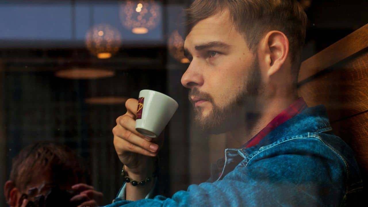 A man with a beard holds a small white cup and looks out a window.