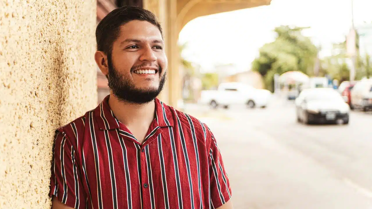 A smiling man in a red striped shirt leans against a textured wall outdoors.