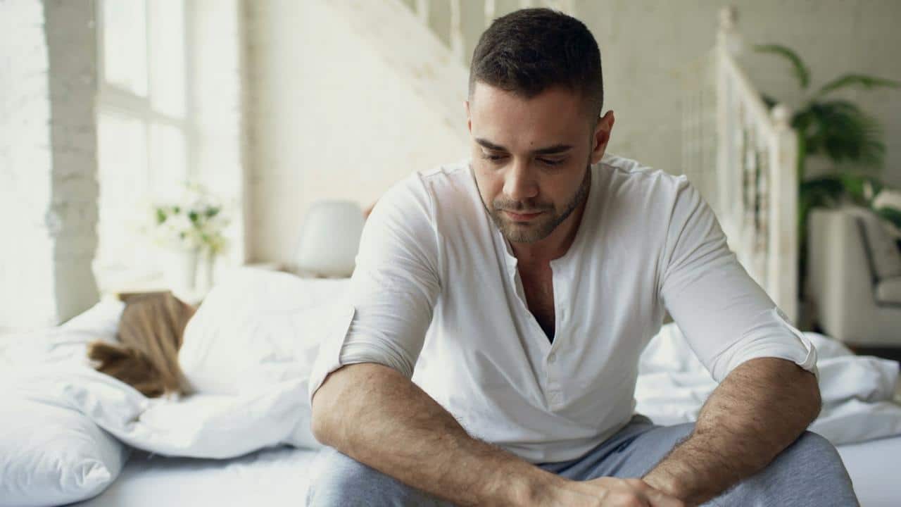 A sad man sitting on the edge of the bed looking down while a woman lies asleep or turned away behind him.
