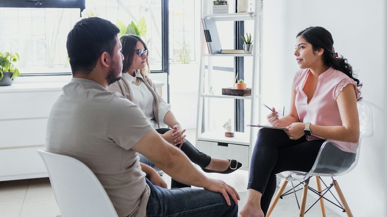 A couple is seated across from a female therapist in a bright, modern office, engaged in a counseling session.