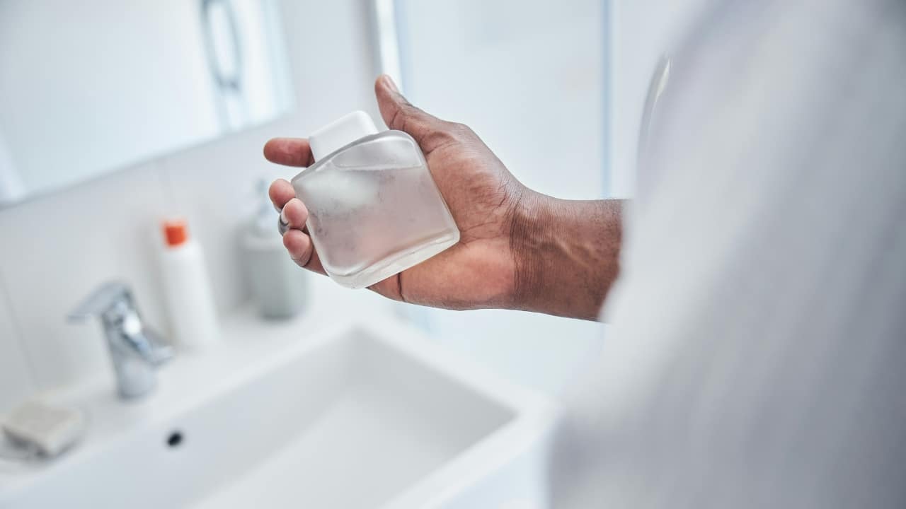 A person's hand holds a frosted glass bottle of liquid over a white bathroom sink.