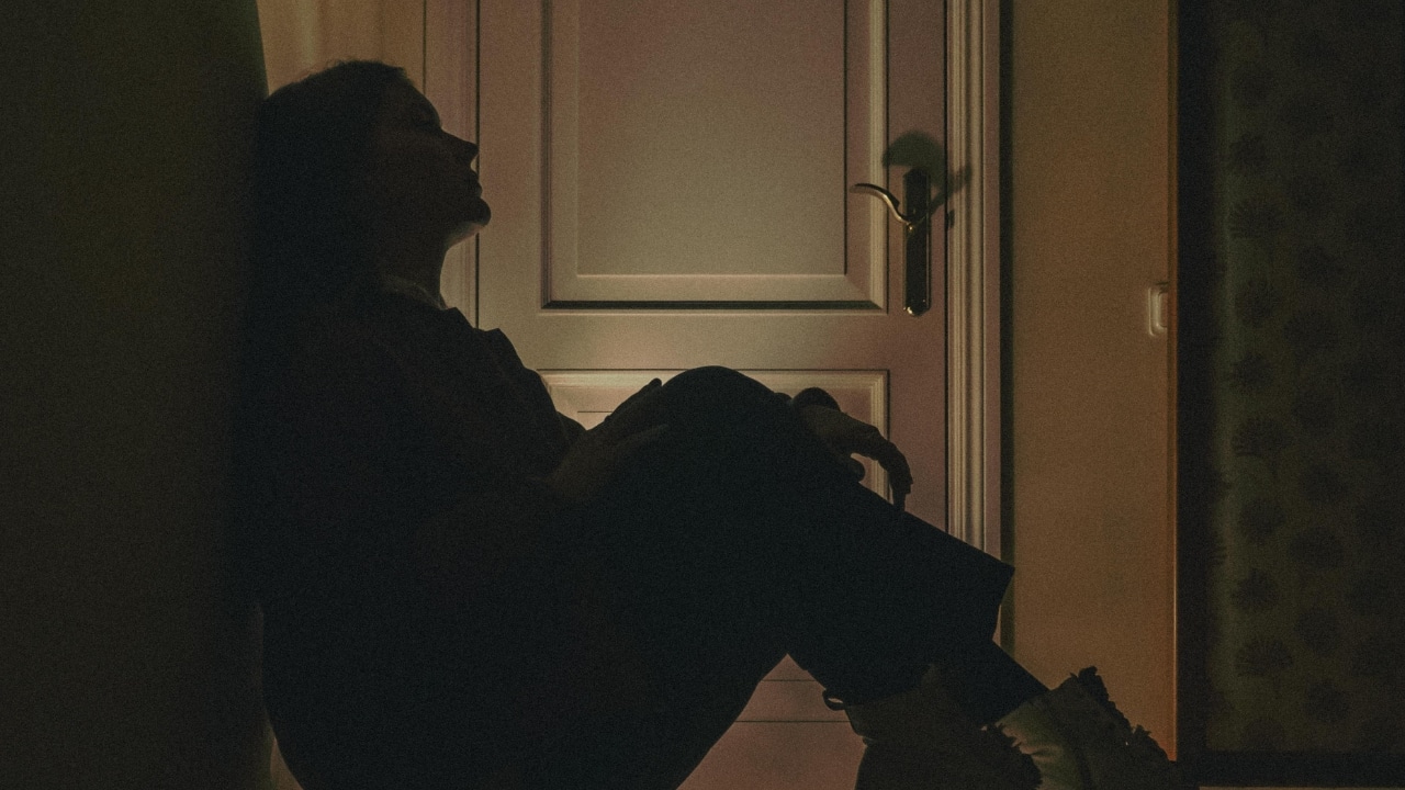 A dark silhouette of a person sits on the floor leaning against a closed door.