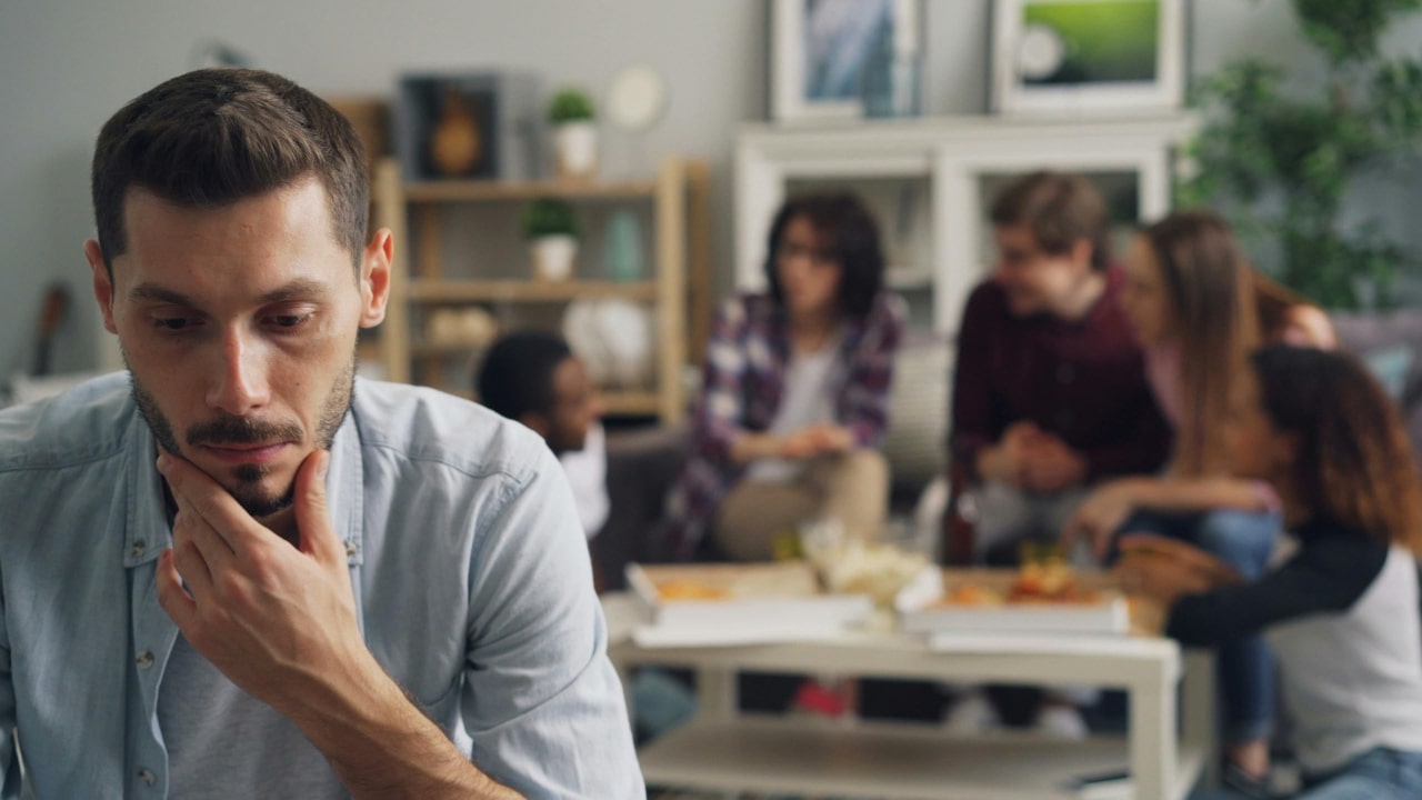 A man looks down pensively while a blurred group of people talks behind him.