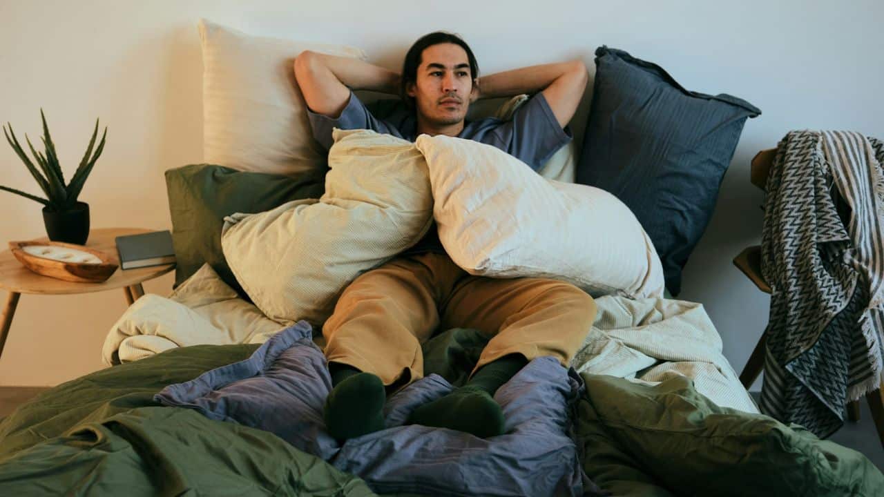A man lies on a bed surrounded by pillows with his hands behind his head.