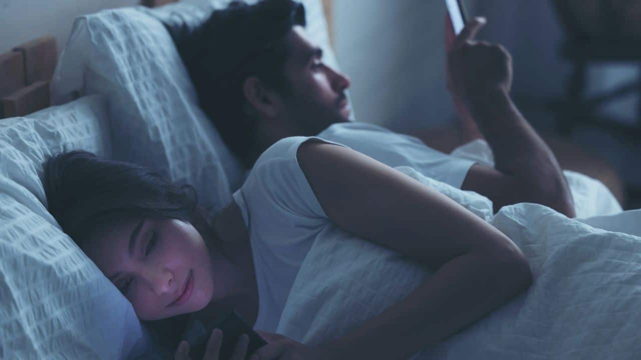 A man and woman lying in bed, facing away from each other while using smartphones.