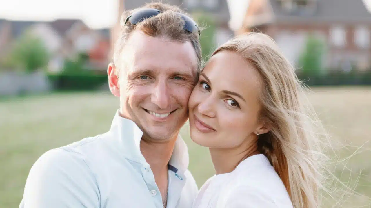 A man and woman pose closely together outdoors while smiling for a direct portrait.
