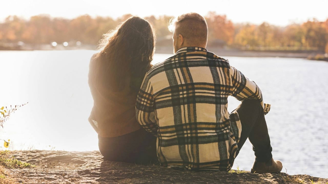 A couple sits on a rocky ledge from behind, looking out over a calm lake.