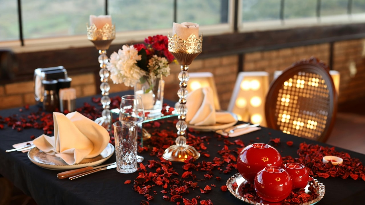 A dining table is decorated with a black cloth, red flower petals, candles, and tall centerpieces.