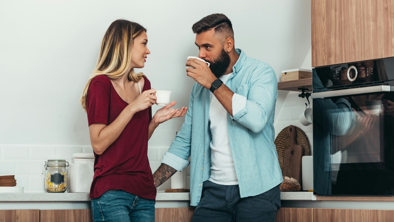 A woman and a bearded man stand in a kitchen while holding white mugs and talking.