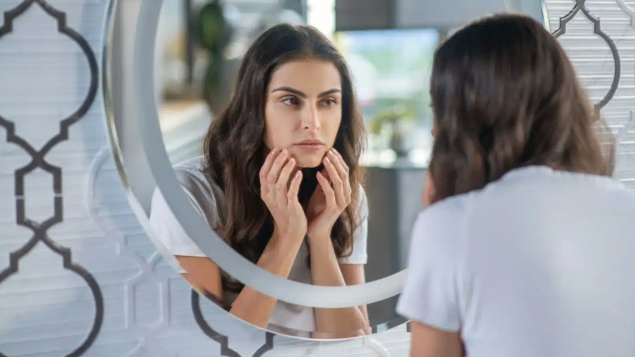 A woman looks at her reflection in a circular mirror while touching her face with both hands.