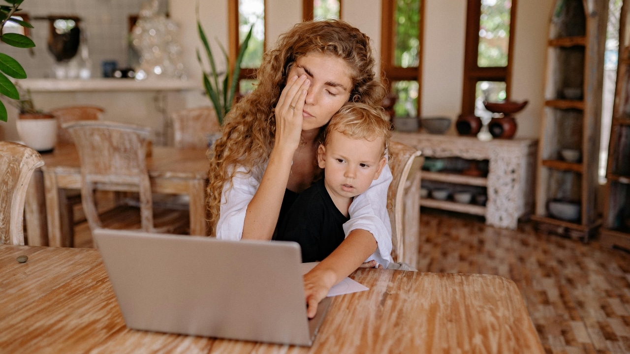 A woman with curly hair sits at a laptop while a small child leans against her arm.