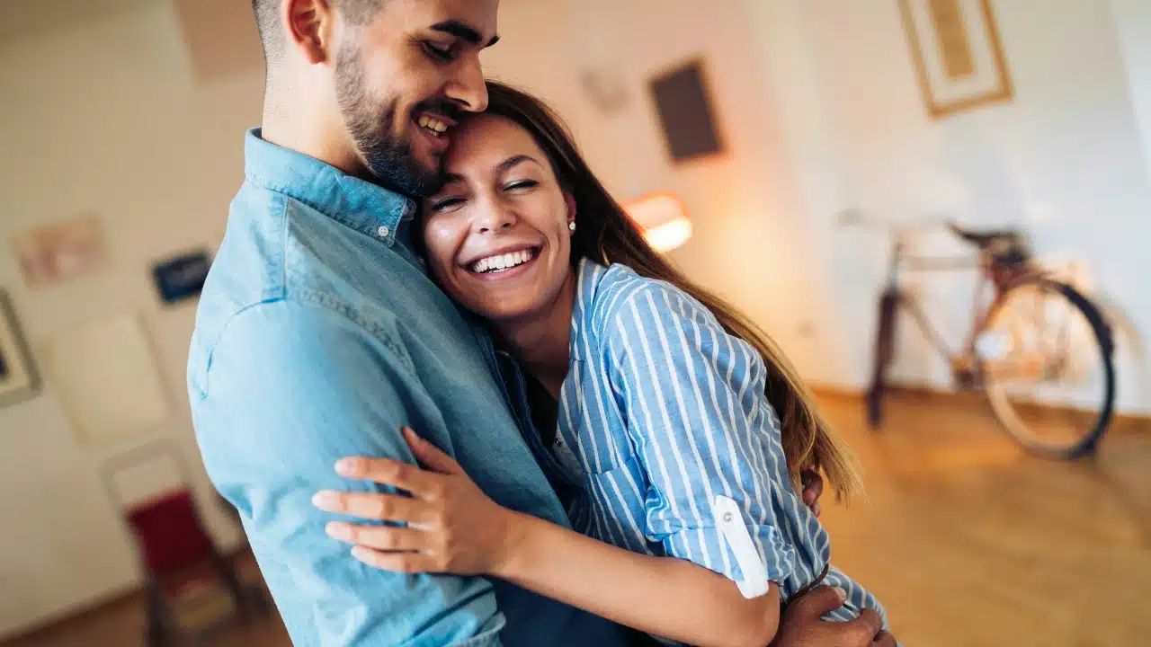 A woman in a striped shirt smiles broadly while embracing a man in a blue shirt.