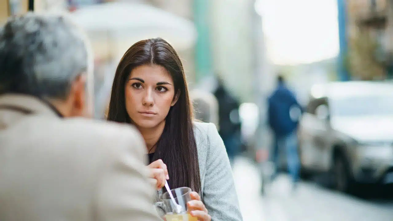 A woman with dark hair looks directly at a man sitting across from her outdoors.