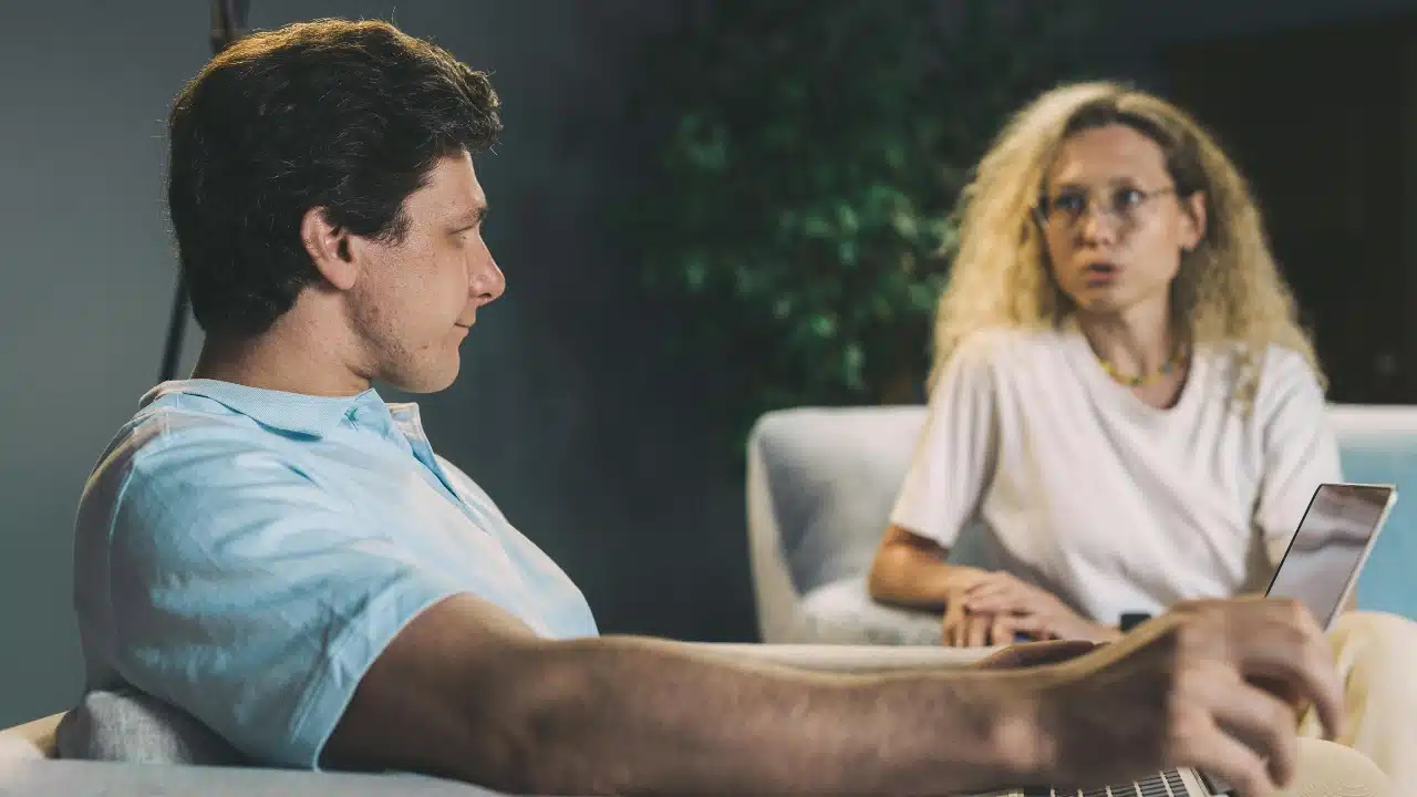 A man in a blue shirt looks at a woman speaking from across the room.