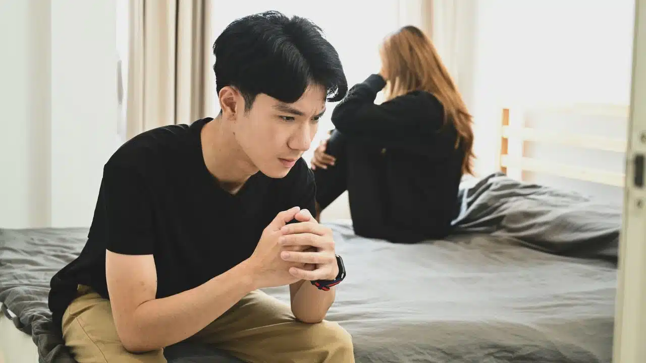 A man sits on a bed with hands clasped while a woman sits facing away.