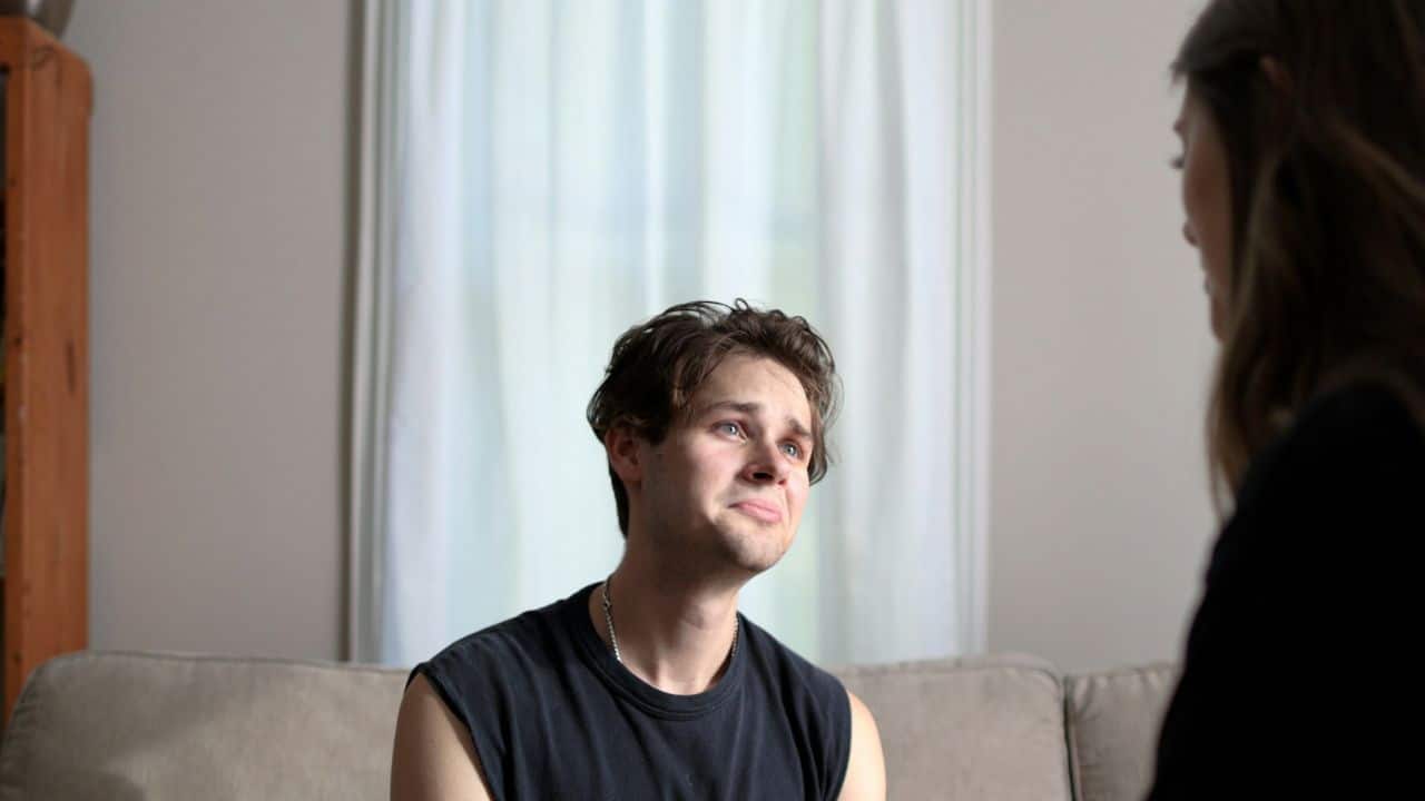 A young man with a sad expression looks up at a woman in a room.