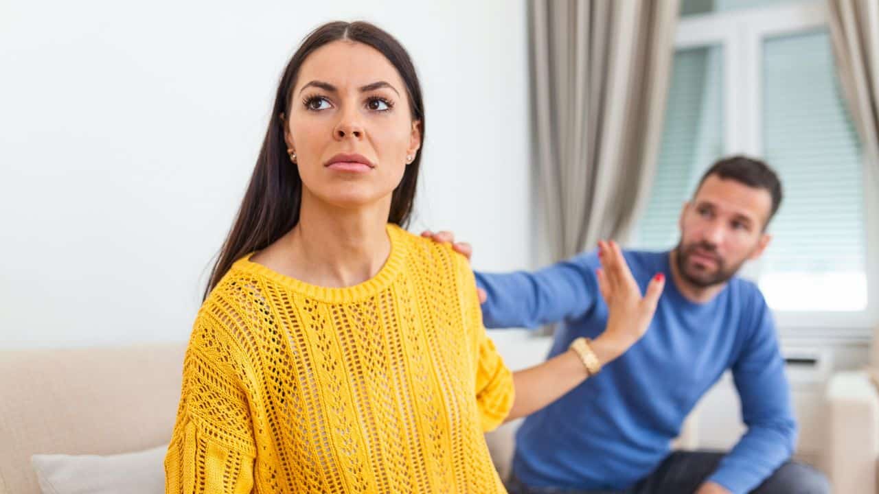 A woman in a yellow sweater looks away while a man behind her reaches out.