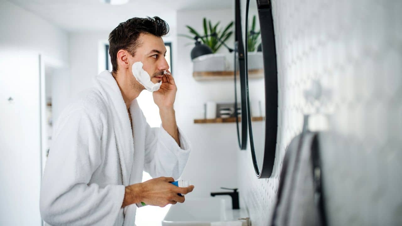 A man in a white bathrobe applies shaving cream to his face before a mirror.