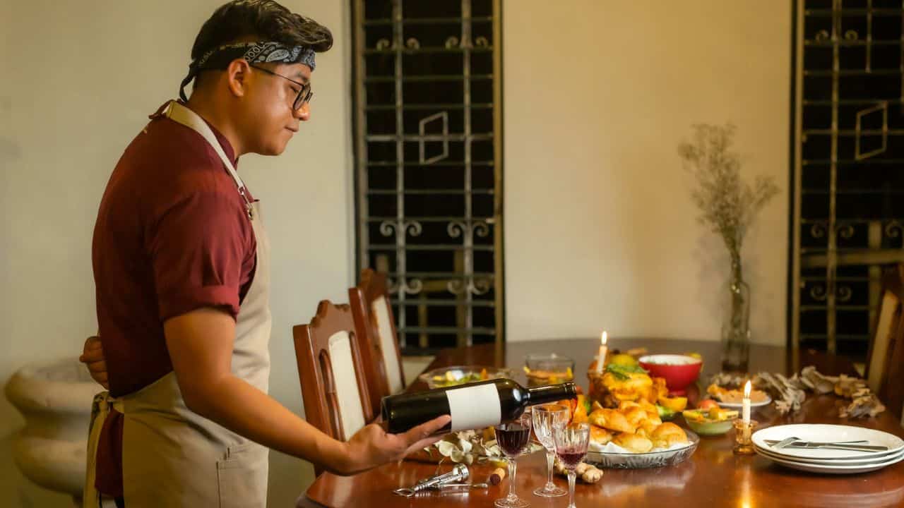 A man in an apron pours wine into a glass at a set dining table.