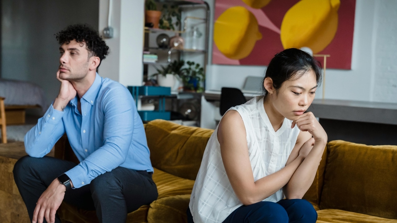 A man and woman sit on a mustard-colored sofa, both looking in different directions.