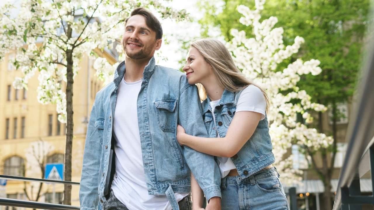 A man and woman in denim jackets walk together outdoors under white flowering trees in bloom.