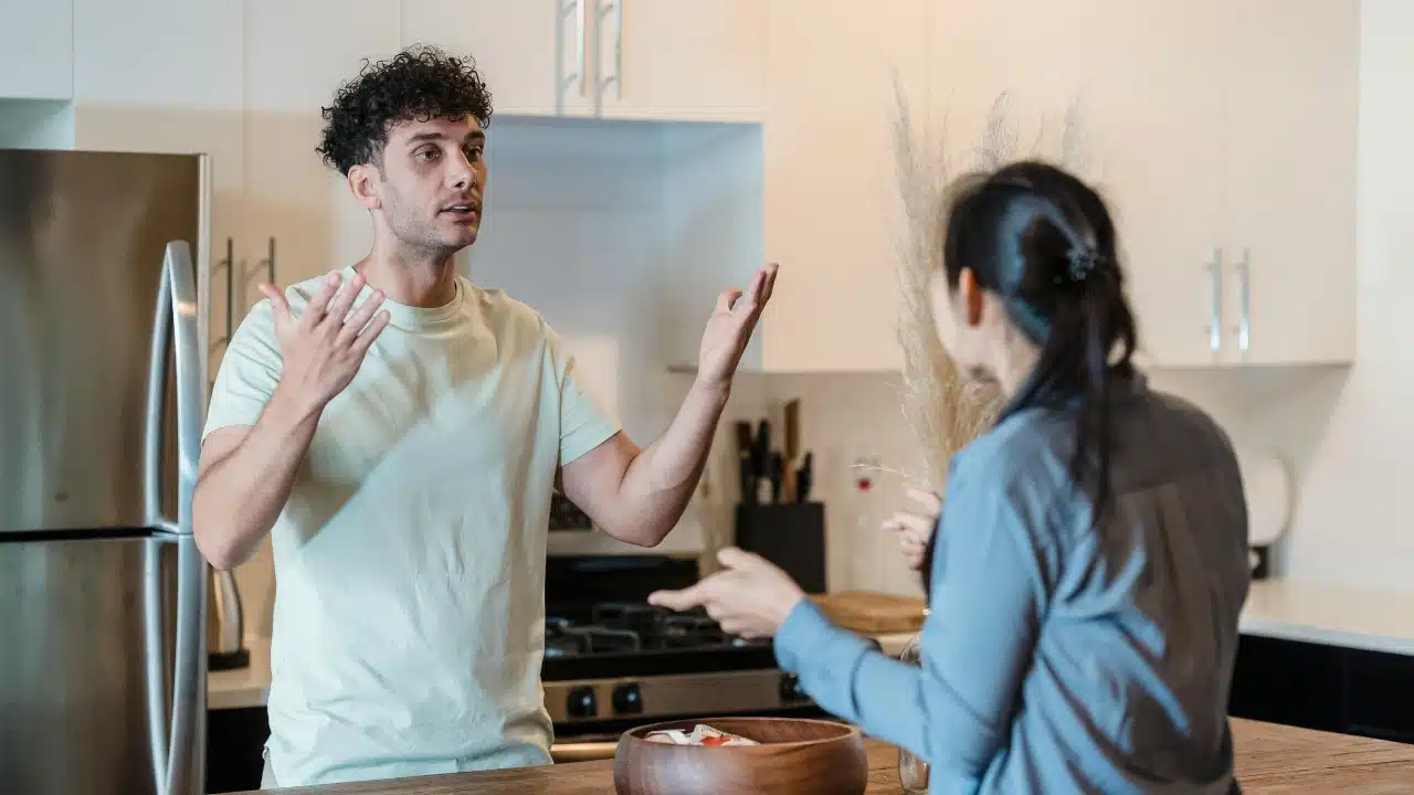 In a kitchen, a man with curly hair raises both hands while speaking to a woman in profile.