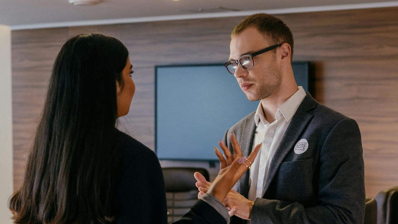 A man wearing a vote button speaks to a woman with long dark hair indoors.