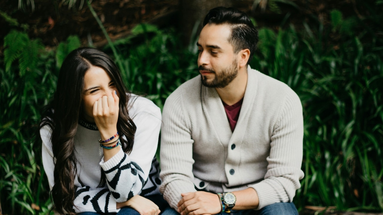 A woman laughs with her hand over her mouth while a man sitting next to her looks on.