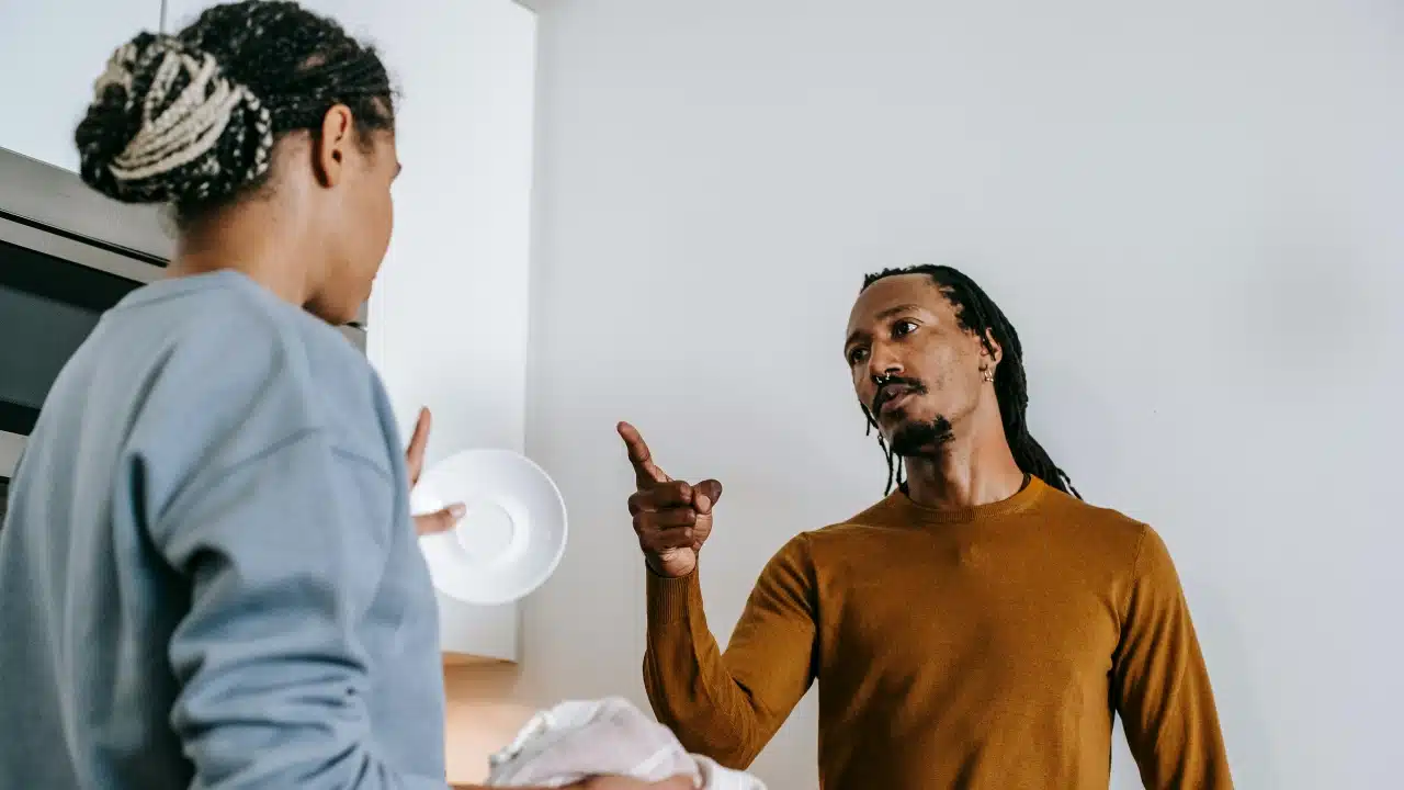 A man with dreadlocks points upward while speaking to a woman holding a white saucer.