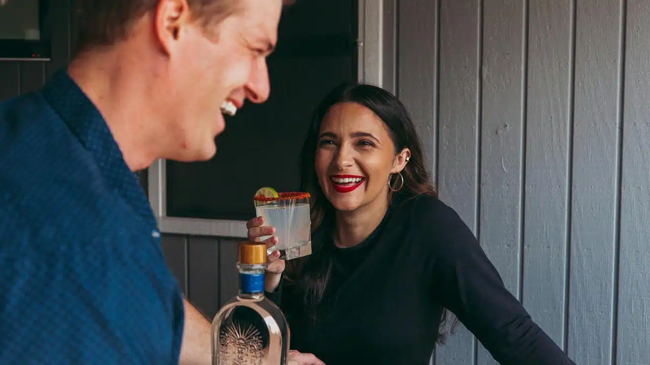 A woman laughing while holding a drink and talking with a man indoors.