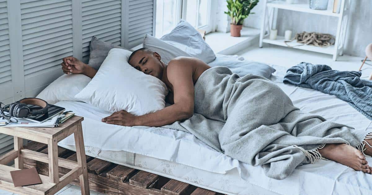 A man sleeping on a bed under a gray blanket in a bright room.
