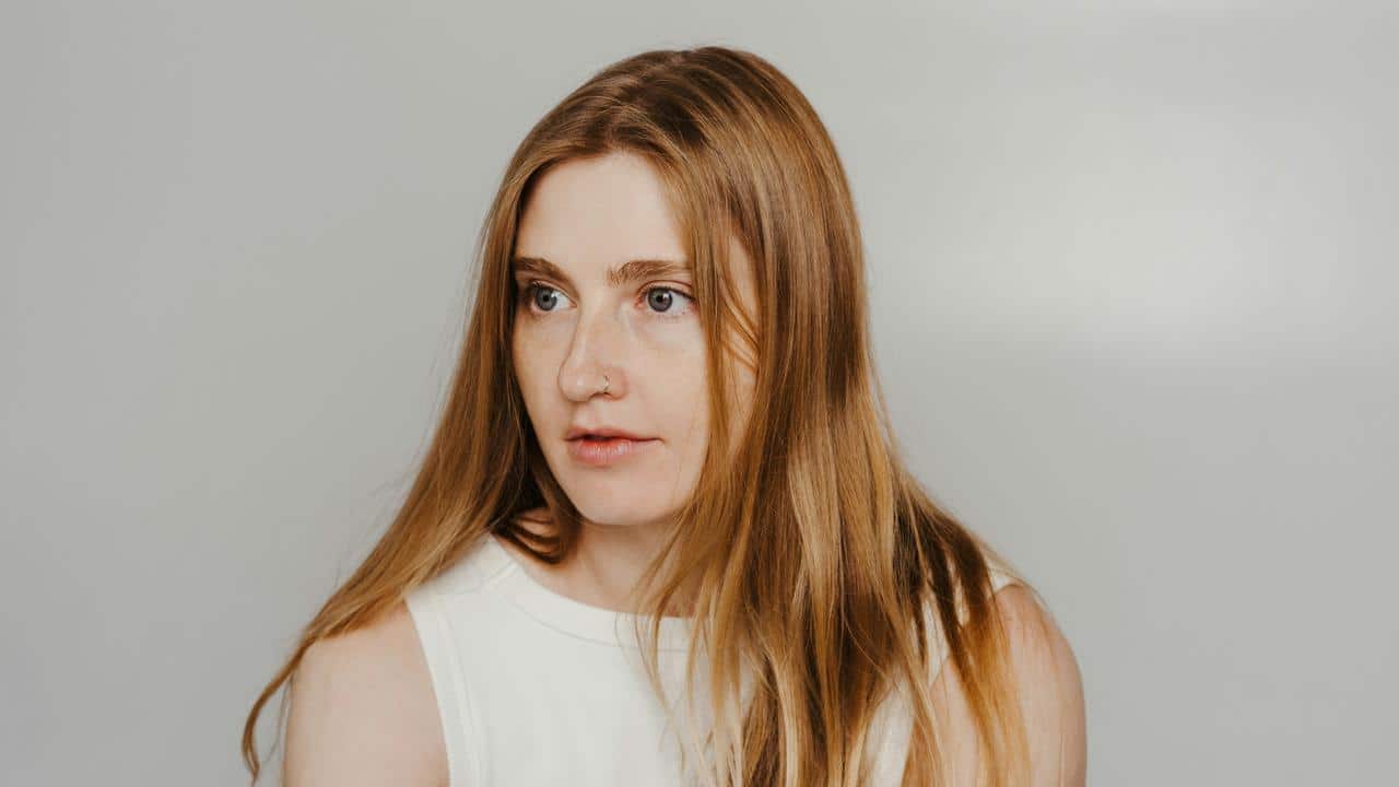 A woman with long red hair looking to the side against a plain background.